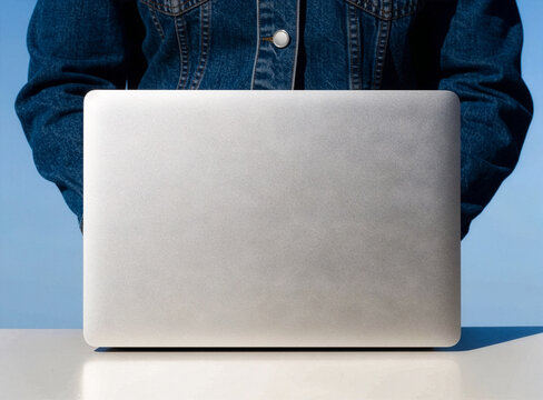 Person in denim jacket behind blank silver laptop against blue sky