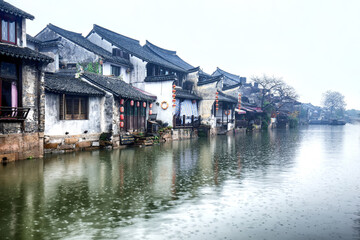 A row of traditional Chinese waterside buildings stands along a calm canal on a misty day, with red lanterns hanging on the facades. © tai