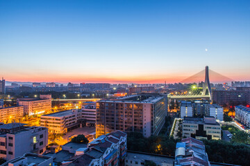 Fototapeta premium A panoramic urban view at dawn, featuring a cable-stayed bridge, lit buildings, and a gradient sky with a visible moon.