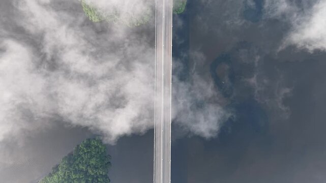 Aerial shot of a narrow bridge with a single car crossing calm water surrounded by mist and green forest, showcasing a tranquil and scenic natural environment at Tasik Raban