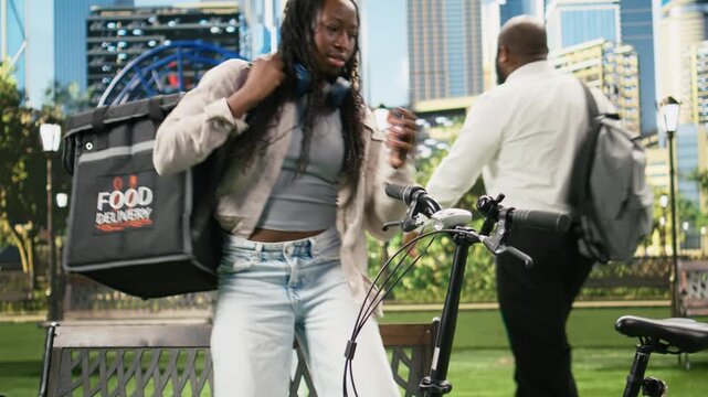 Smiling courier looks at the camera while seated with a smartphone, checking an on-demand mobile app for a new order with a thermal backpack nearby.