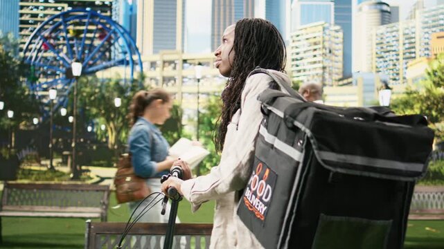 Woman bicycle courier stands with a bike and insulated delivery backpack in a city park, ready to continue work after checking the next route.