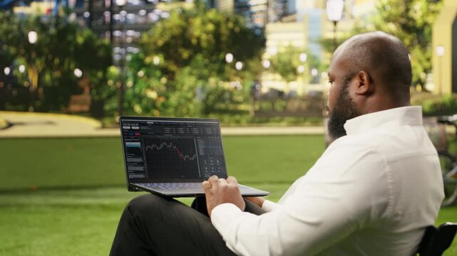Man reads financial data graphs on a forex web platform using a laptop with candlestick charts and market analysis trends in a sunny green park, illustrating foreign exchange investing.
