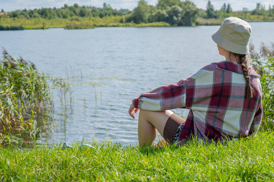 Woman in a light sun hat and checked shirt sits sideways on bright green grass beside the water with her knees tucked underneath. She looks toward a forested shore, creating a scene of leisure
