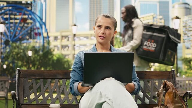 Focused woman studies content on a laptop while sitting on a wooden bench in an urban park, surrounded by greenery and modern buildings during everyday outdoor technology use in a city setting.