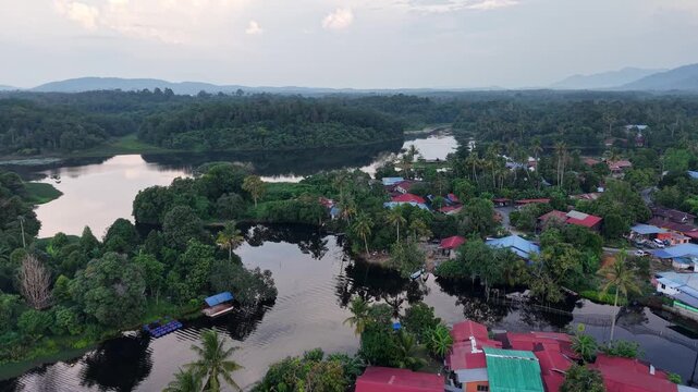 Aerial view of colorful houses along a calm river surrounded by lush green forest and distant hills under a cloudy sky in a peaceful rural setting at Tasik Raban