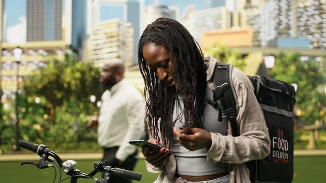 Anxious woman courier fidgets while waiting for a new order request, repeatedly checking a delivery mobile app beside a bicycle and thermal bag in a city park during gig work outdoors.