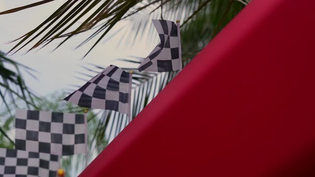 Small checkered racing flags waving in the wind outdoors near palm trees and red banner