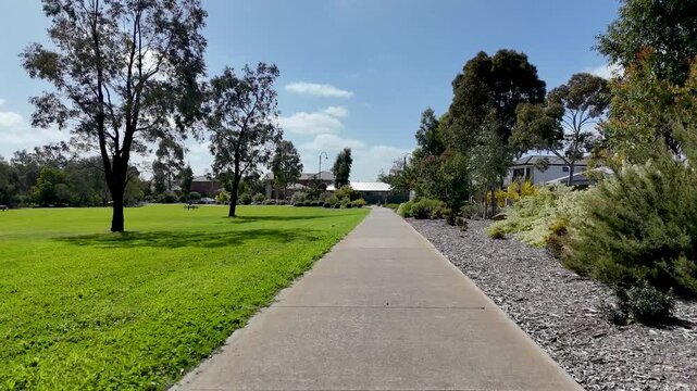 A concrete pedestrian footpath in a suburban park, featuring lush green lawns, native gum trees, landscaped garden beds, and modern housing bordering the open reserve. Tarneit, Melbourne, Australia