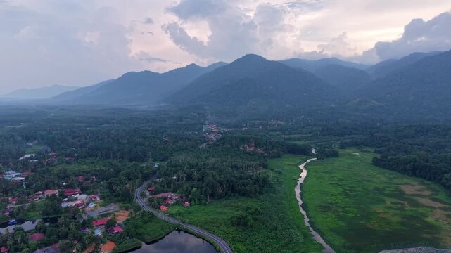 Aerial view of a winding road passing through a lush green valley surrounded by dense forest and towering mountains under a cloudy sky at dusk at Tasik Raban