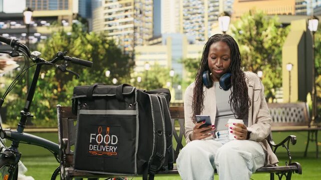 Concerned woman courier checks a smartphone with coffee in hand while resting beside a delivery bag in an urban park, waiting anxiously for a new order.