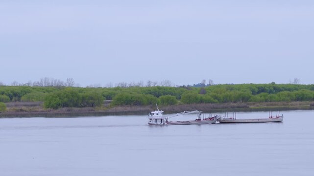 Small cargo boat towing a barge along a wide river against a backdrop of dense green trees
