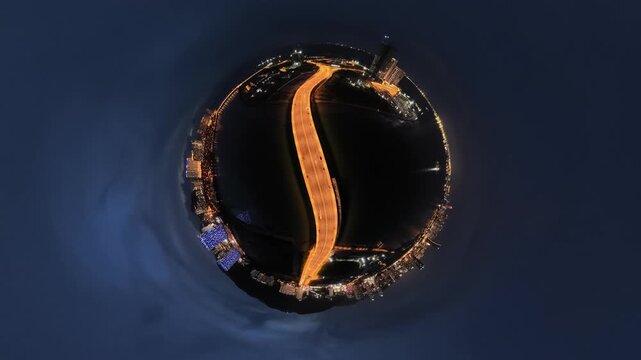 An aerial tiny planet view capturing an illuminated curved bridge spanning a river in a bustling city at night at Gurney Bay