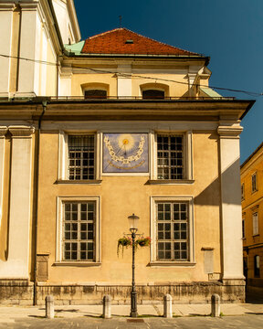 A sunlit historic building wall featuring a traditional sundial mounted between upper windows, displaying Roman numerals and a Latin inscription as a decorative timekeeping feature.