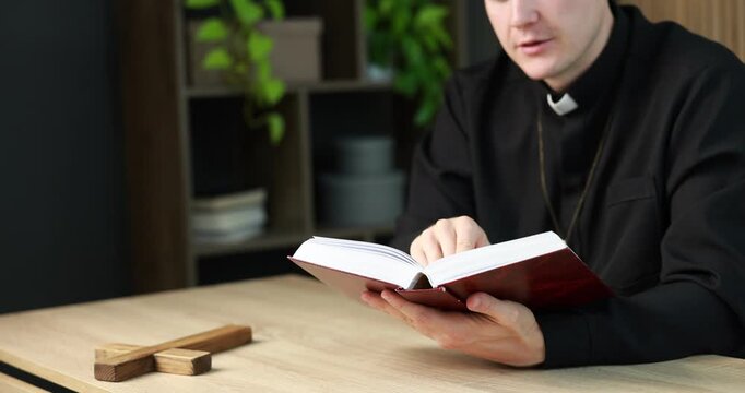 Priest in cassock reading Holy Bible at wooden table indoors, closeup