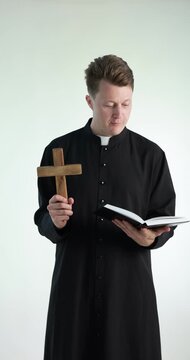 Priest in cassock with wooden cross reading Holy Bible on white background