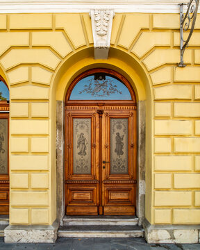 An elegant wooden double door featuring etched glass figures sits within a striking yellow stone arched entrance, highlighting the intricate craftsmanship and classical beauty of the building facade.