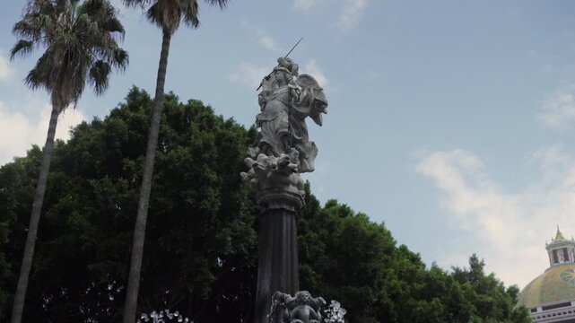 Archangel San Miguel fountain in central zocalo with Puebla Cathedral towers in background framed by green trees on cloudy day