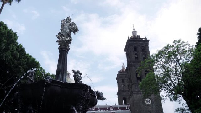 Archangel San Miguel fountain in central zocalo with Puebla Cathedral towers in background framed by green trees on cloudy day