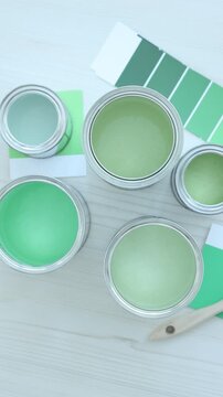 Metal cans with paint in shades of green, brush and color palettes on white wooden table, flat lay. Camera spinning