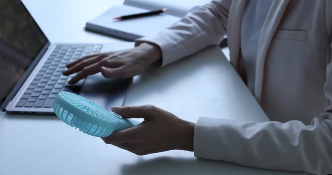 Woman with mini fan working on laptop at white desk in office, closeup