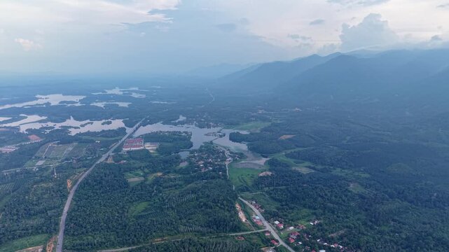 Aerial scenery of a peaceful rural area featuring lush green forest, calm lake, winding roads, small village houses, and mountains under a cloudy sky at Tasik Raban