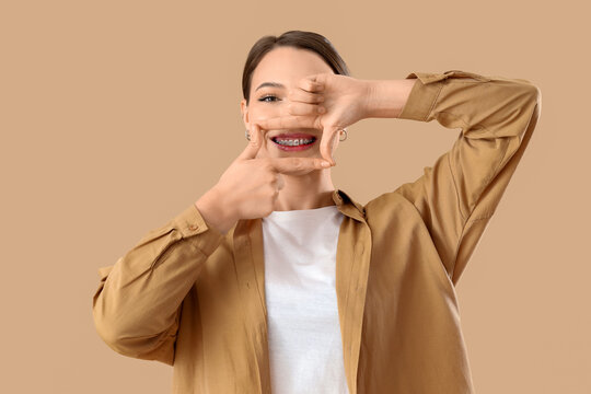 Smiling young woman with dental braces making frame gesture on beige background