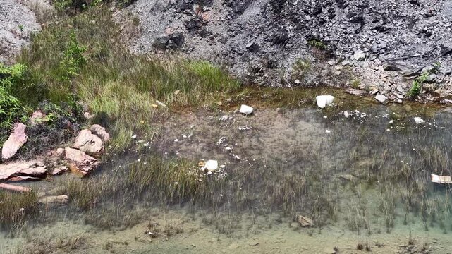 High angle drone image of rocky quarry shoreline with shallow water, marsh grass and scattered litter.