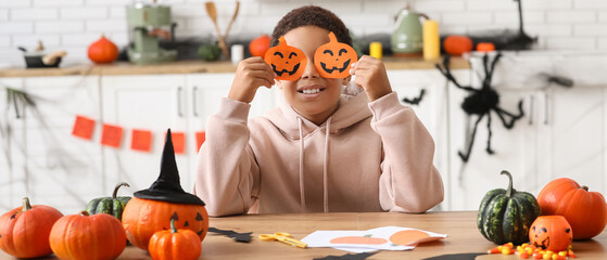 Naklejka premium Halloween celebration and childhood joy, happy boy holding paper pumpkin masks over eyes while crafting at home kitchen table with autumn decorations