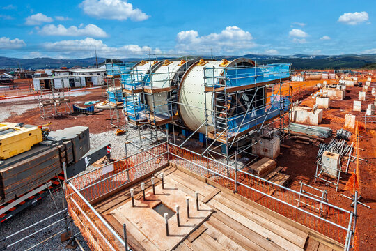 Large-scale iron ore mining infrastructure construction site in Brazil featuring a massive industrial mill, scaffolding, heavy machinery, and red soil.