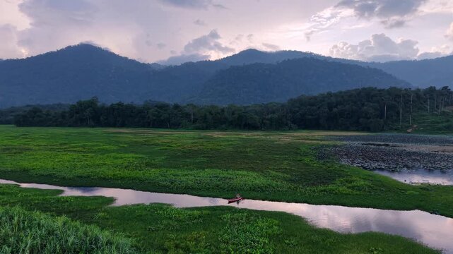 A tranquil river winds through vibrant green fields with dense forest and towering mountain range under a cloudy sky at dusk, showcasing peaceful natural beauty at Tasik Raban