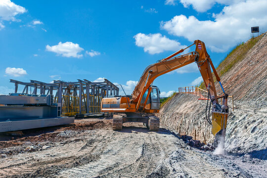 Excavator with hydraulic breaker breaking rock at a construction site. Large steel beams and structural framework visible under a blue sky.