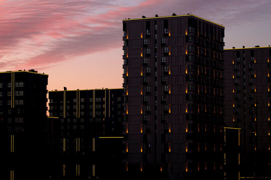 Night city background with illuminated lights on modern architecture &mdash; real estate concept. Beautiful atmospheric urban sunset sky with pink colorful clouds over dark residential block exterior.