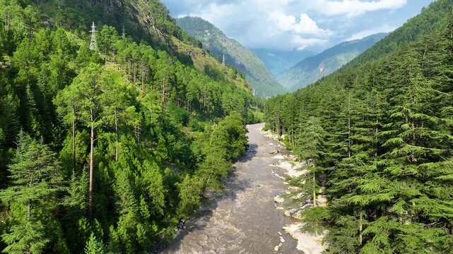 Drone orbit of sunlit coniferous mountain ridges and summer monsoon clouds hanging over the winding Parvati River in the Indian Himalayas.