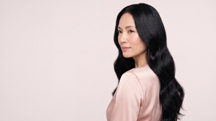 Obraz premium Asian woman with long dark wavy hair wearing a pink silk top stands against a plain neutral background in a studio portrait.