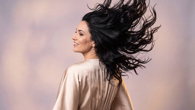 Brunette woman wearing a silky robe turns around with a joyful expression while her voluminous dark hair dynamically tosses in the air creating a dramatic motion effect.