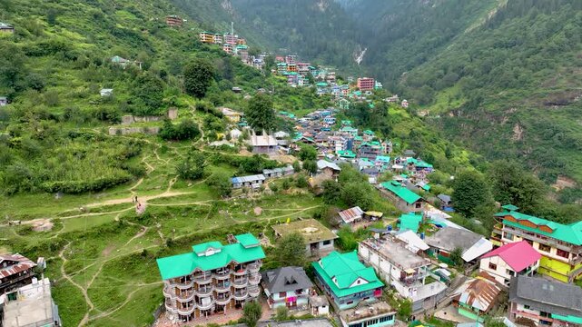 Aerial tracking shot over the colorful tin roofs and lush agricultural terraces of Barshaini village, a vibrant trekking basecamp in the Parvati Valley of Himachal Pradesh, India.