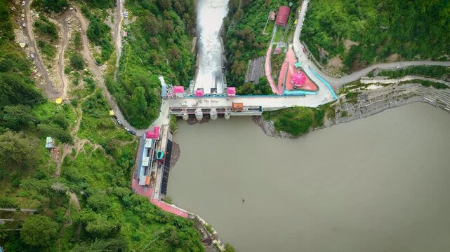 Top-down aerial shot of churning white water discharging from the concrete spillway of Barshaini dam, highlighting hydroelectric infrastructure in Parvati Valley, India.