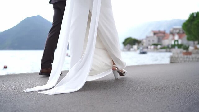 Bride and groom standing on waterfront promenade in Perast Montenegro, wedding dress with slit and mountain bay view, low angle video