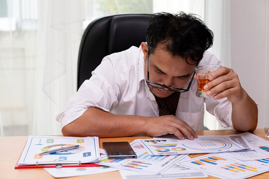 Stressed adult man drinking alcohol at office desk with work documents and glasses, showing fatigue, stress, exhaustion and emotional pressure during difficult business work