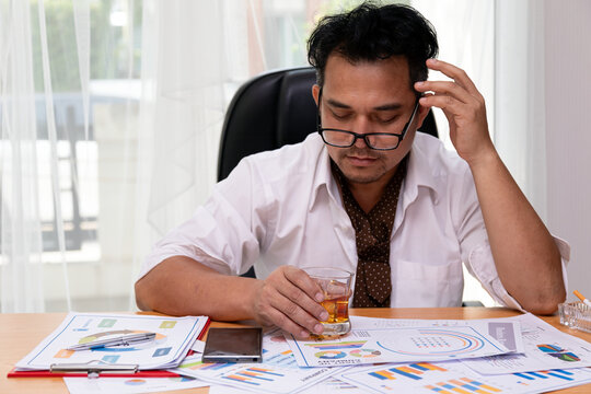Stressed adult man drinking alcohol at office desk with documents, glasses and work reports, showing fatigue, pressure and emotional strain