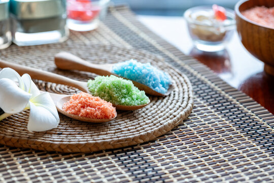 Colorful bath salt crystals in wooden spoons with white frangipani flower on woven mat for spa, wellness, relaxation, beauty, therapy and natural body care