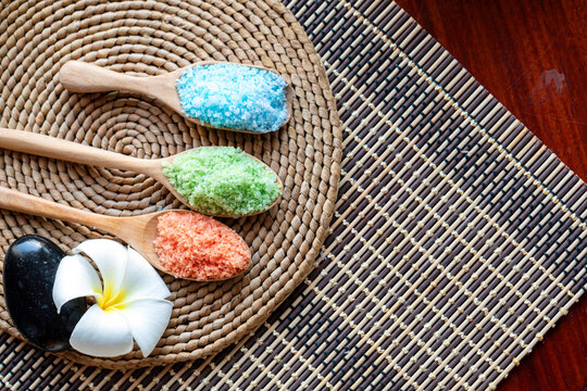 Colorful bath salt in wooden spoons with black spa stone and white tropical flower on woven mat for wellness, relaxation, beauty, therapy and natural body care