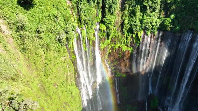 Aerial view of tall multi-stream waterfall cascading down lush green cliffs, rainbow visible