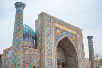 Ornate facade of Sher-Dor Madrasa at Registan Square in Samarkand, Uzbekistan, featuring turquoise domes, slender minarets, monumental pointed arches, intricate geometric mosaic tilework © pploylp