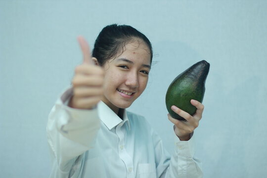 Smiling young woman holding a large avocado and giving a thumbs up gesture, representing healthy food and positive lifestyle choices.