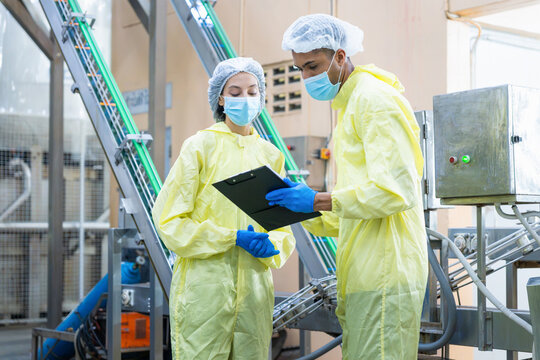 Food factory workers in protective suits and face masks inspecting production line with clipboard, Quality control inspectors wearing hairnets and sterile clothing checking process in plant