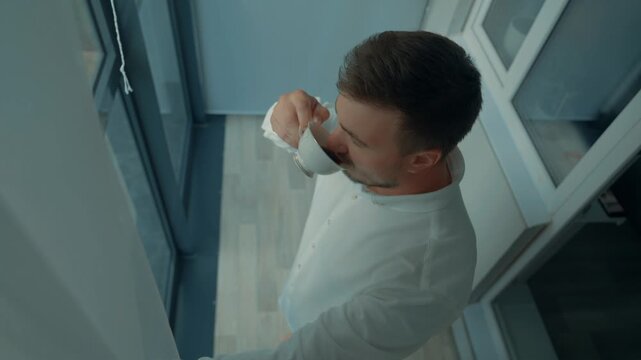 A young man is looking out the panoramic window and drinking coffee.