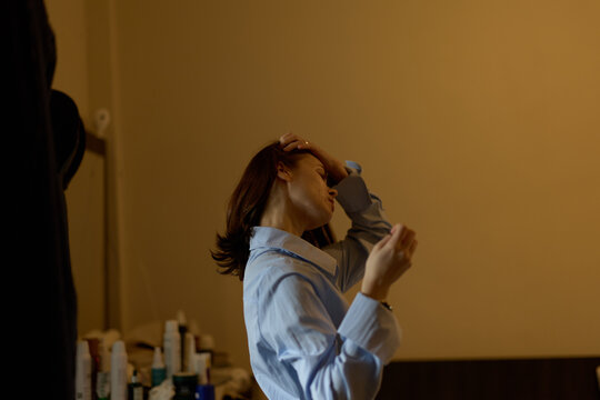 A woman in a blue shirt adjusts her hair while standing indoors near a table with bottles in a softly lit room, capturing a quiet personal moment.