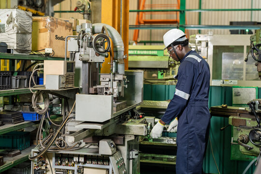 African adult male operator stands beside machine station and watches work area closely, supervising setup position and process readiness for precision production task in industrial workshop.
